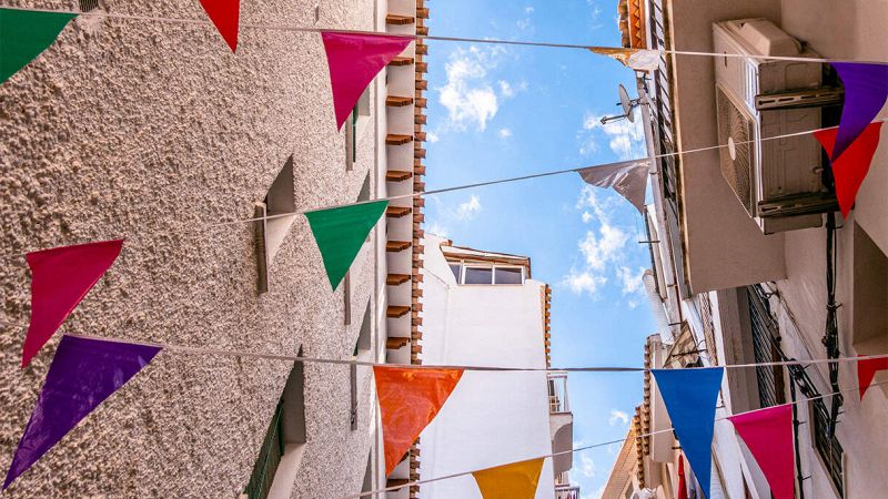 Bandiere triangolari colorate appese tra case di una stretta via, sotto un cielo azzurro con poche nuvole