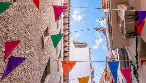 Bandiere triangolari colorate appese tra case di una stretta via, sotto un cielo azzurro con poche nuvole