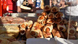 Banco di sagra con vassoi pieni di würstel avvolti in pasta di pane cotti al forno, in primo piano.