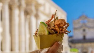 Cono di carta con fritto misto di pesce e spicchio di limone in primo piano, sullo sfondo edifici storici sfocati