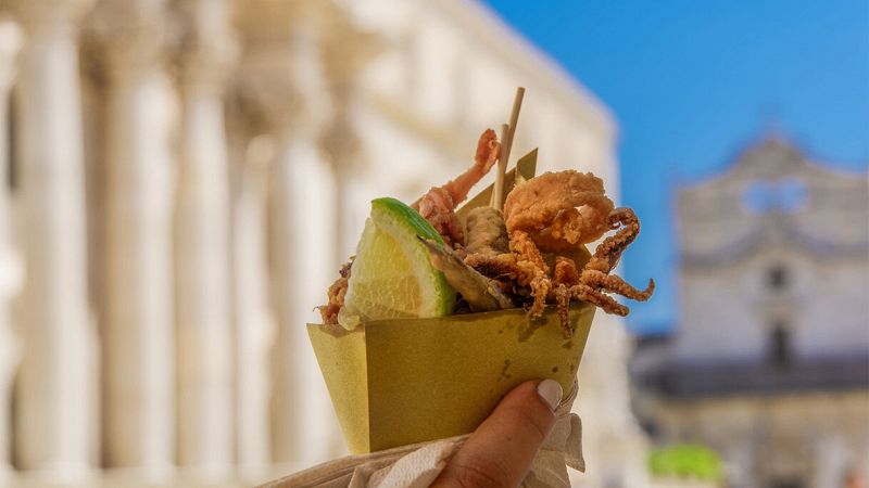 Cono di carta con fritto misto di pesce e spicchio di limone in primo piano, sullo sfondo edifici storici sfocati