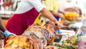 Grande porchetta arrostita su banco di sagra, con addetti al taglio e spezie fresche in primo piano