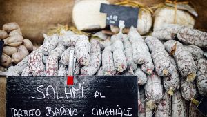 Banco di mercato con salami artigianali impilati e lavagna con scritta “salami al tartufo, Barolo, cinghiale”