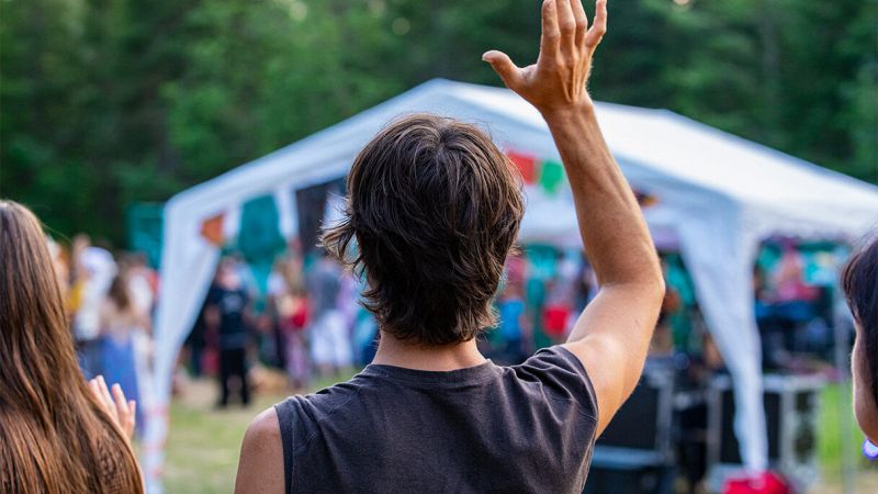 Ragazzo di spalle alza una mano tra la folla durante una festa all’aperto con tendone bianco e bandierine colorate
