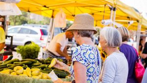 Due donne anziane scelgono zucchine e verdure fresche a un banco di mercato all’aperto con tende gialle