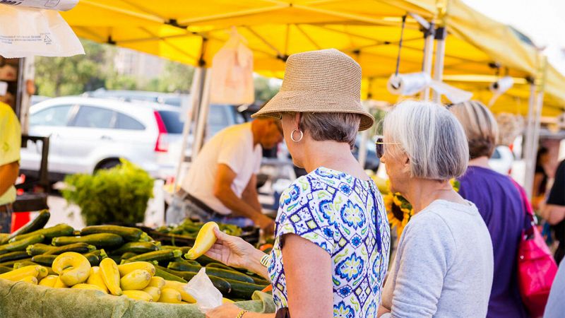 Due donne anziane scelgono zucchine e verdure fresche a un banco di mercato all’aperto con tende gialle