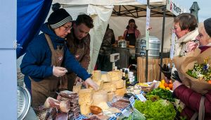 Bancarella di mercato con formaggi, salumi, verdure e fiori; venditori servono clienti infreddoliti sotto il tendone