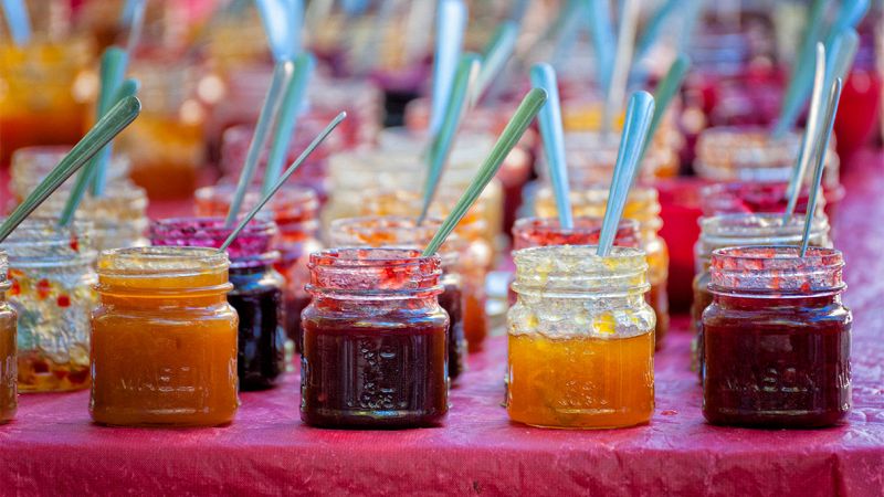 Barattoli di marmellate colorate allineati su un banco, ciascuno con un cucchiaino di metallo inserito