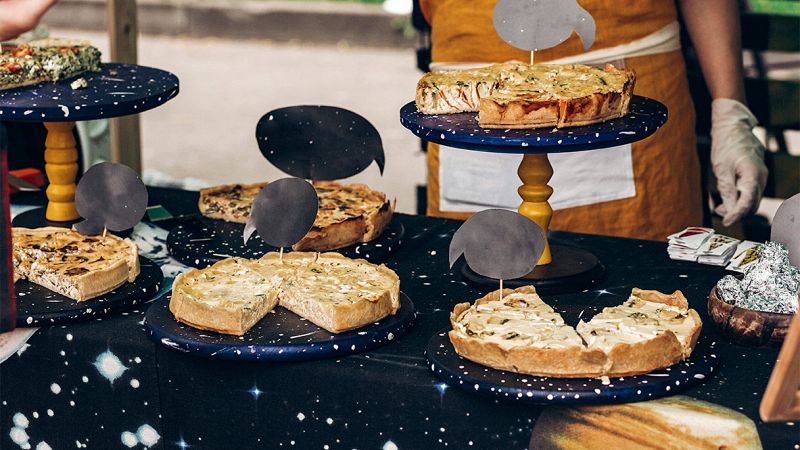Banco di sagra con diverse torte salate esposte su alzatine blu, etichette nere e persona con grembiule sullo sfondo