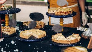 Banco di sagra con diverse torte salate esposte su alzatine blu, etichette nere e persona con grembiule sullo sfondo
