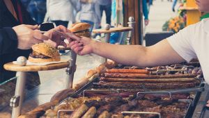 Bancarella di street food con salsicce e carne alla griglia; cliente paga un panino al venditore
