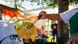 Mano regge con una pinza una grande focaccia fritta dorata in una sagra all’aperto, con persone sedute sullo sfondo