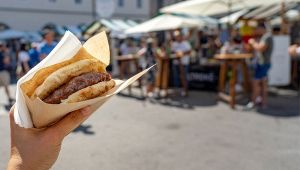 Panino con salsiccia tenuto in mano in primo piano, sullo sfondo sfocato bancarelle e persone a una festa all’aperto