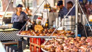 Grande griglia sospesa con cosce di carne arrosto a una sagra, con cuochi al lavoro sullo sfondo sfocato