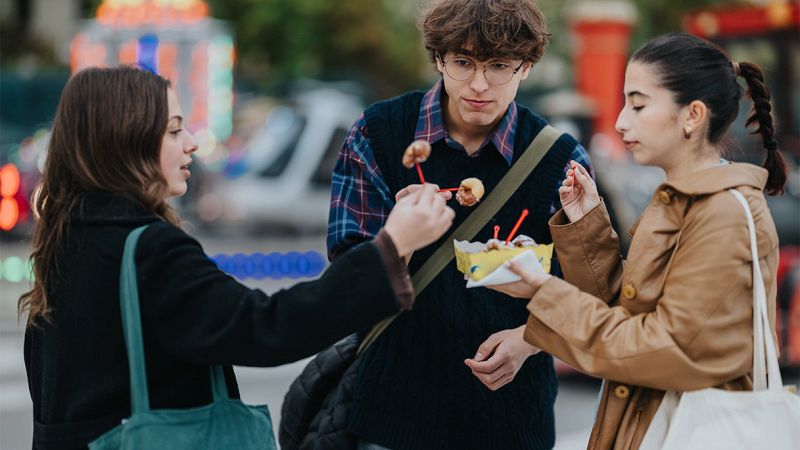 Tre giovani all’aperto condividono uno spuntino su stecchini durante una festa con luci colorate sullo sfondo