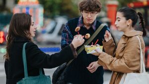 Tre giovani all’aperto condividono uno spuntino su stecchini durante una festa con luci colorate sullo sfondo