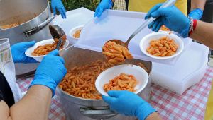 Volontari con guanti blu servono penne al pomodoro da una grande pentola in piatti di plastica a una sagra