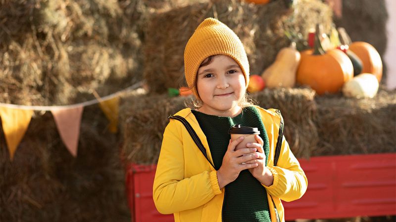 Bambina con berretto giallo tiene una bevanda calda davanti a balle di fieno decorate con zucche e bandierine alla sagra