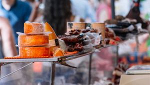 Banco di mercato con forme di formaggio arancione e salumi confezionati sottovuoto, persone sfocate sullo sfondo