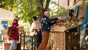 Persone al mercato all’aperto comprano frutta e verdura da un banco decorato con trecce d’aglio sotto gli alberi