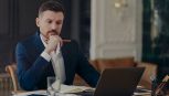 Serious male entrepreneur holds chin makes notes in diary works on laptop computer makes money online dressed formally writes information. Confident managing director involved in working process