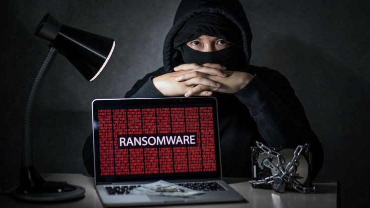 Un hacker incappucciato seduto su una scrivania con un computer davanti che espone la scritta in rosso ransomware