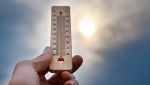 Una mano tiene un termometro che segna oltre 30°C, con il sole sfocato e un cielo nuvoloso sullo sfondo, a indicare il caldo intenso