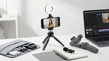 Smartphone montato su treppiede con ring light accesa e microfono esterno, posizionato su una scrivania moderna da content creator.