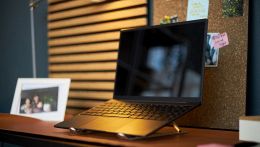 Laptop nero ultra-sottile posizionato su un supporto rialzato in alluminio, su una scrivania da home office con bacheca in sughero e lampada.