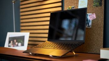 Laptop nero ultra-sottile posizionato su un supporto rialzato in alluminio, su una scrivania da home office con bacheca in sughero e lampada.