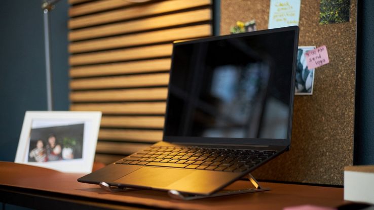 Laptop nero ultra-sottile posizionato su un supporto rialzato in alluminio, su una scrivania da home office con bacheca in sughero e lampada.