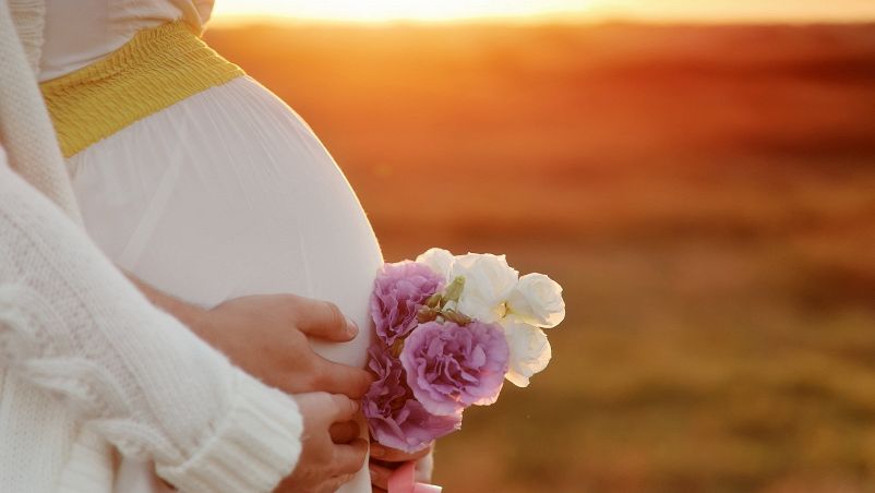 Al tramonto, profilo del ventre di una donna in gravidanza con mani poggiate che stringono un mazzo di fiori