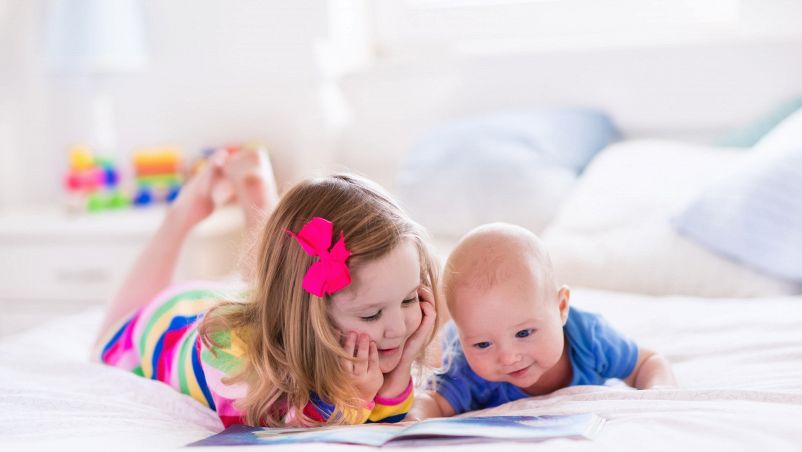 Bambina sorridente che legge un libro e gioca nel letto con il fratellino neonato. Bambini in stanza luminosa
