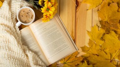 Su tavolo in legno, visti dall’alto, da sx a dx, un libro aperto, una coperta di lana color avorio, fiori gialli finti, foglie marroncine autunnali