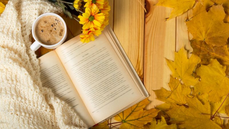Su tavolo in legno, visti dall’alto, da sx a dx, un libro aperto, una coperta di lana color avorio, fiori gialli finti, foglie marroncine autunnali