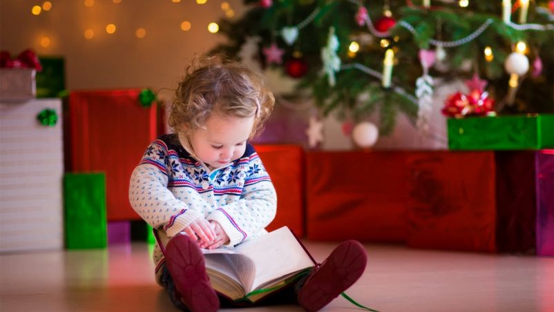 Una bimbetta impegnata a leggere sul pavimento di casa sua, con alle spalle un albero di Natale illuminato e regali impacchettati dalle carte lucenti