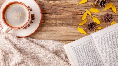 Vista dall’alto di: tazza di caffè, foglie ingiallite, pigne, libro aperto a metà, coperta bianca fatta a maglia, tutto appoggiato su tavolo in legno.