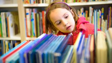 Una bimba castana con giacca rossa sta decidendo quale albo illustrato prendere in biblioteca; vista frontale; sfondo sfocato con altri libri