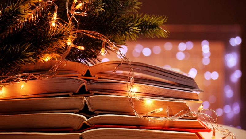 Sulla sinistra in basso una colonna di libri aperti appoggiati gli uni sugli altri, in alto albero di Natale; sulla destra luci non messe a fuoco