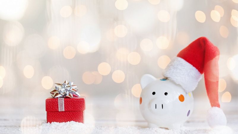 In ptimo piano un salvadanaio bianco a forma di maialino con cappello natalizio e pacco rosso con fiocco argento; sfondo chiaro con lucine gialle, sfocate