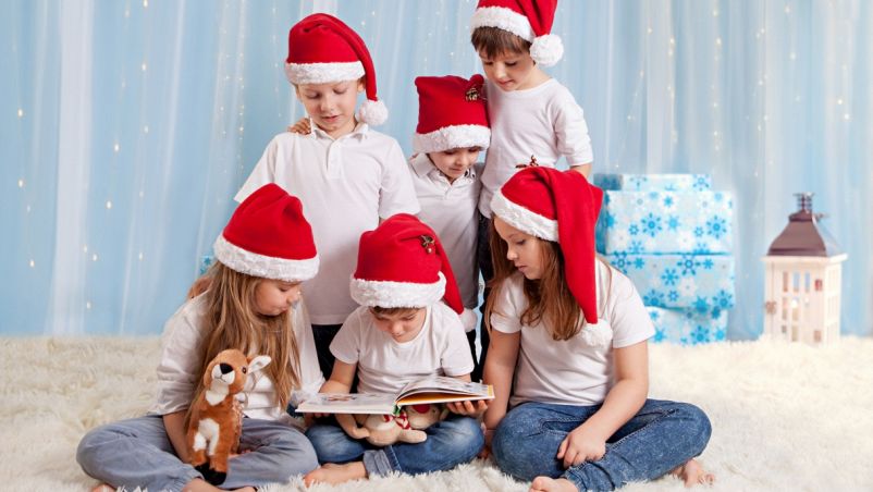 Tre bambini in piedi e tre seduti davanti, quello in mezzo con un libro in mano; indossano cappelli natalizi in uno studio fotografico e guardano interessati il volume