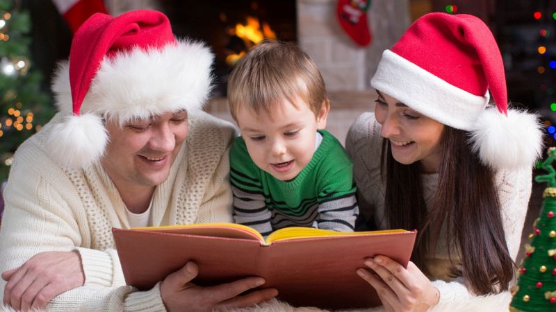 Tra i genitori con un cappello da Santa Claus in testa, un bimbo di 3 anni sta leggendo un libro coricato per terra; sullo sfondo un caminetto acceso