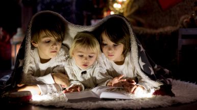 Da sx a dx un fratello di 5 anni, un fratellino di 3 e una sorella di 4 sfogliano un libro distesi a terra; luci natalizie gialle sfocate dietro di loro