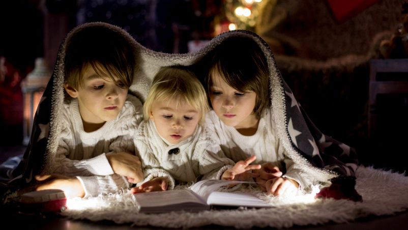 Da sx a dx un fratello di 5 anni, un fratellino di 3 e una sorella di 4 sfogliano un libro distesi a terra; luci natalizie gialle sfocate dietro di loro