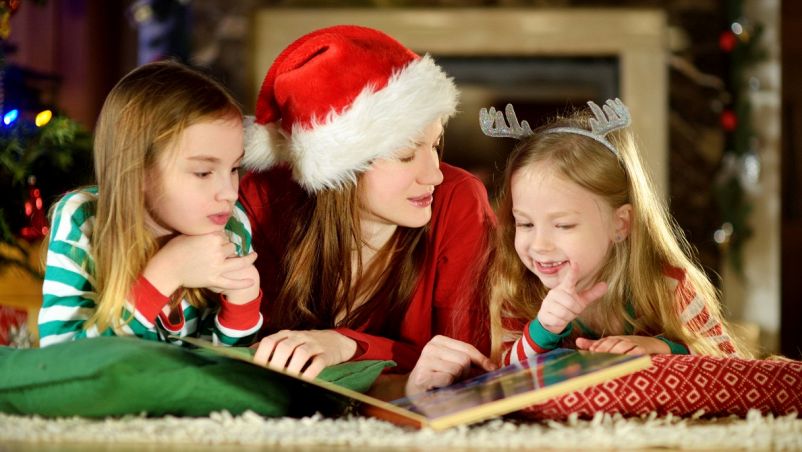 Mamma con cappello natalizio, figlia minore con cerchietto da renna e figlia maggiore con abiti verdi e rossi leggono coricate un libro dal grande formato