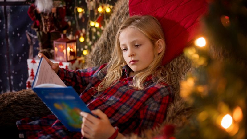 Una bambina di 7 anni immersa nella lettura su una poltrona accogliente, con luci soffuse; intorno atmosfera natalizia e abete illuminato