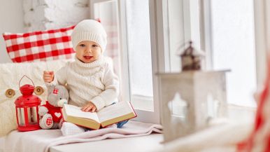 Sulla dx sfocata una lanterna; sx un bimbo di 1 anno con un grande libro in mano, con abbigliamento invernale; siede sul davanzale; fuori c'è la neve