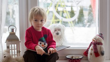 Bambino di 3 anni con maglione rosso e libro in mano, su un davanzale con una lanterna, un peluche a forma di pupazzo di neve e una ciotola con cioccolatini