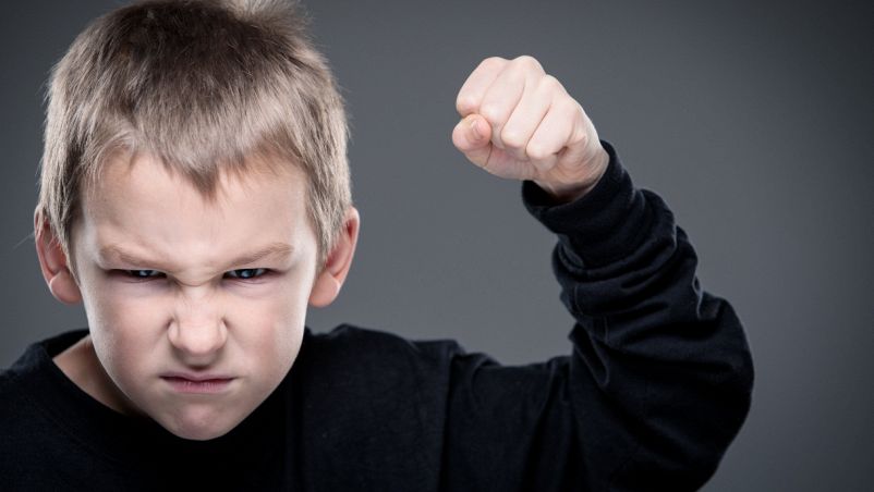 Espressione arrabbiata, occhi aggrottati e bocca corrucciata: un bambino biondo con maglia nera di 4 anni alza il pugno sinistro in segno di sfida