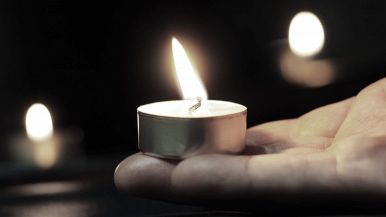 In primo piano la fiamma di un lume posato sul palmo di una mano, sullo sfondo luci di candele sfocate: Giornata della Memoria, commemorazione della Shoah